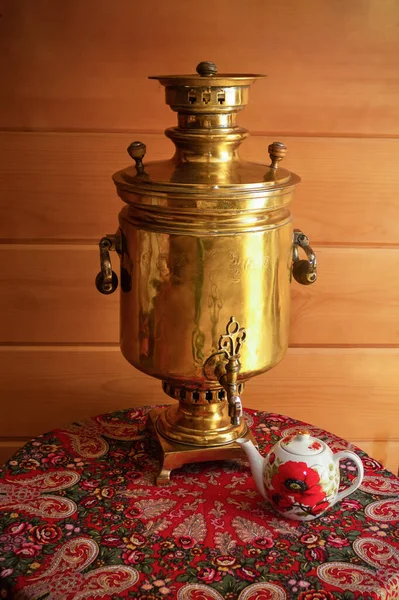 Yellow traditional Russian samovar on table with variegated red tablecloth and teapot. Against background of wooden plank wall.