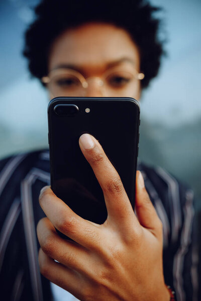 4k. Charming African American woman holding her phone looks in camera. Businesswoman or student. Close up.
