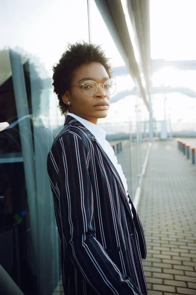 4k. Perjalanan, Digital. Charming African American woman in elegant striped suit. Berdiri dekat dinding kaca dan terlihat di kamera. Pebisnis wanita atau mahasiswa. Pemikiran. Langit. Stok Lukisan  
