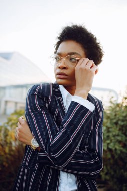 4 bin. Seyahat et, Dijital. Zarif çizgili takım elbiseli çekici Afrikalı Amerikalı kadın, sol tarafına bakıyor, gözlüklerine dokunuyor. Arka planda duruyor. Havaalanının yakınındaki çalılıklar. İş kadını ya da model. Bahçe