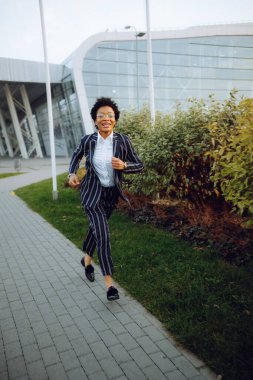 4 bin. Seyahat et, Dijital. Zarif çizgili takım elbiseli çekici bir Afrikalı Amerikalı kadın kameraya bakıyor, koşuyor, gülümsüyor ve kendini harika hissediyor. Havaalanının yakınındaki çalılıklara doğru koştu. İş kadını.