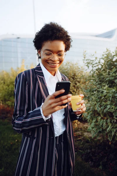4k. Perjalanan, Digital. Wanita Afrika Amerika yang menawan dengan pakaian bergaris elegan dengan telepon dan memegang secangkir kopi. Berdiri melawan semak-semak. Pebisnis wanita atau model. Dan melihat di telepon dan tersenyum. Taman Stok Gambar