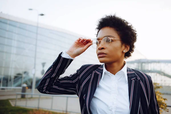 4k 입니다. 여행, 디지털. 우아 한 줄무늬 옷을 입은 아프리카계 미국인 여성이 왼쪽을 보고는 안경을 만진다. 공항 근처 덤불 뒤에 멈춰 있어. 비지니스 우먼 또는 모델. 텃밭 로열티 프리 스톡 이미지
