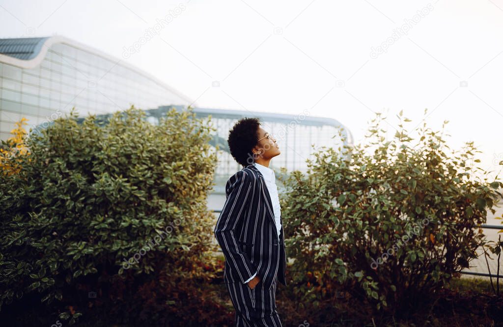 4k. Travel, Digital. A charming African American woman in an elegant striped suit, looks at the sky, and waiting for her flight. She stands on background of bushes near the airport. Businesswoman or