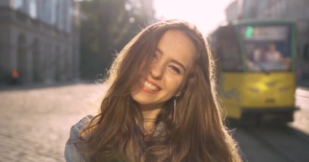 Uzun kıvırcık sarı saçlı çekici kız kameraya bakar ve gülümser. Arka planda tramvay gezileri. — Stok video