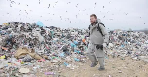 Er is een enorme zwerm vogels boven de stortplaats. een man in een overall met een gasmasker in zijn hand loopt langs de stortplaats — Stockvideo