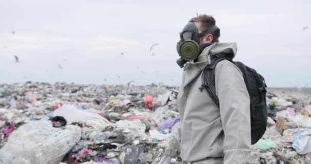 Symboliskt skott miljöskydd, man med gas mask på deponi, titta runt, titta på kamera, sopspår passerar genom deponi i bakgrunden — Stockvideo