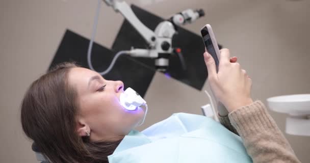 Femme assise sur une chaise de dentiste. Alors que le blanchiment des dents, elle navigue sur le réseau social Instagram sur son smartphone — Video