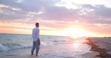 İyi yapılı genç ve çekici bir adam denizin kenarında duruyor ve güzel gün batımını seyrediyor. Pembe gökyüzü