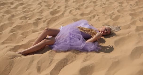 穿着浅紫色衣服的迷人的金发女子躺在沙滩上，手里拿着一束铃铛。女孩躺在沙漠的沙地上 — 图库视频影像