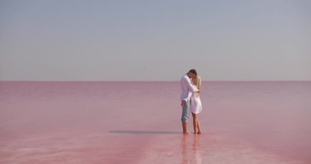 Feliz pareja romántica encantadora de pie sobre un hermoso fondo de lago mineral rosa parece desierto. andsome hombre abrazando sonriente bonita rubia y besarla — Vídeo de stock