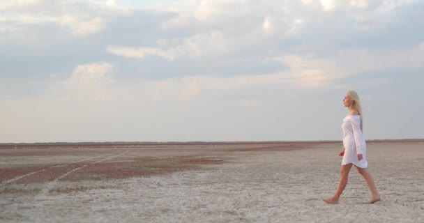 Full length shot of woman walking barefoot σε κυματιστή άμμο σε αργή κίνηση. Νεαρή κυρία με λευκό φόρεμα περνάει τις διακοπές της στο σαφάρι στην έρημο. Γυναίκα τουρίστρια ψάχνει γύρω από άγριο τοπίο. — Αρχείο Βίντεο