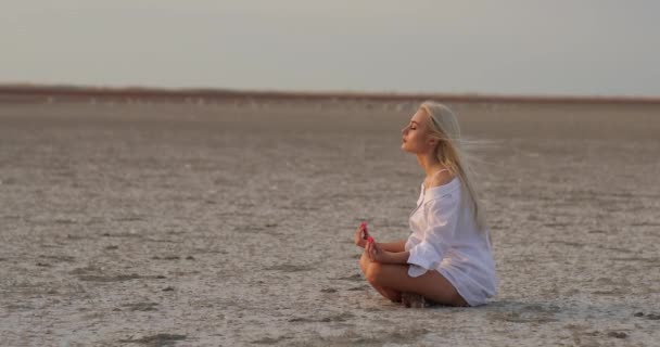 Uma jovem atraente sentada na areia. Meditação. Mulher de ioga à beira-mar relaxante em uma postura de ioga de lótus zen despreocupada na praia ao nascer do sol. Mulher meditando — Vídeo de Stock