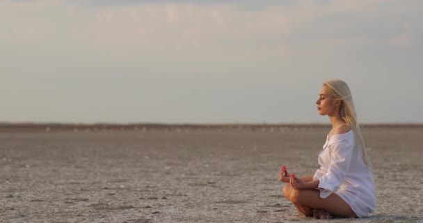 Egy fiatal nő jógázik a parton. Egy fiatal nő meditál a Lotus Pose-ban desszertként. Lazíts a meditáción. Fiatal kaukázusi nő gyakorolja jóga szabadban a természet. — Stock videók