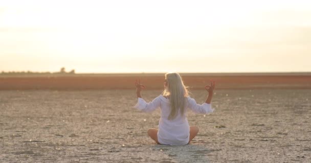 Вид спереду медитативної молодої жінки, що сидить у позі лотоса і кладе руки на коліно в положенні Om, роздумуючи надворі вранці на світанку, на фоні сонця — стокове відео