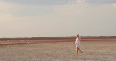 Beyaz elbiseli güzel sarışın kadın yürüyor. Uçan saçlı çekici bir manken. Güzel kız saçlarını sallıyor. Yavaş çekim.