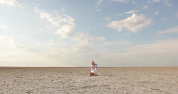 Légi felvétel. Vonzó fiatal nő szőke hajjal, fehér ruhában, a homokon ülve. A háttérben sok sirály sétál a homokon.. — Stock videók