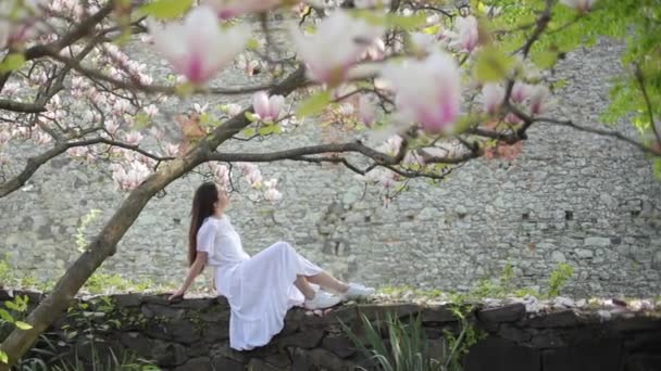 Mooie jonge stijlvolle vrouw zit en houdt magnolia bloem buiten in het stadspark. Romantisch teder meisje in casual trendy kleding en bril tegen park. — Stockvideo