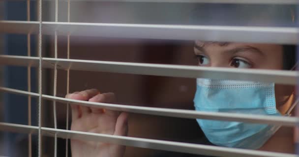 El chico con una máscara médica mira por la ventana a través de las persianas. Cuarentena durante una epidemia. Un niño con una máscara médica mira por la ventana. Autoaislamiento en cuarentena, coronavirus, covid 19 — Vídeos de Stock