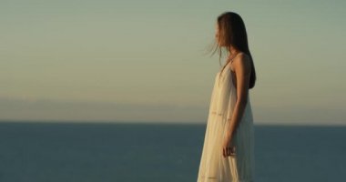 Beyaz elbiseli güzel bir kadın uçurumun kenarında duruyor ve günbatımının tadını çıkarıyor. Okyanus ufkunun arka planında. 4K video. Rüzgarda çırpınan elbise. Rüzgâr saçlara esiyor.