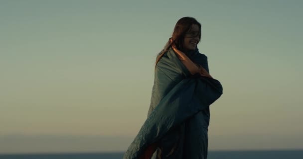 Een mooie jonge vrouw in groene deken staat op de rand van een klif en geniet van de zonsondergang. Tegen de achtergrond van de oceaanhorizon. 4K video. Lachende mooie vrouw. Aangenaam humeur. — Stockvideo
