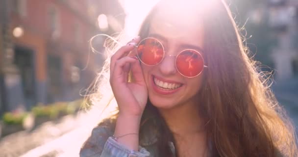 Mujer joven en gafas de sol redondas de color rosa mira a la cámara y sonríe. — Vídeos de Stock