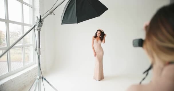 モデルはスタジオでポーズ。女性写真家は白い背景に立ってピンクの夜のドレスで女性の写真を撮る — ストック動画