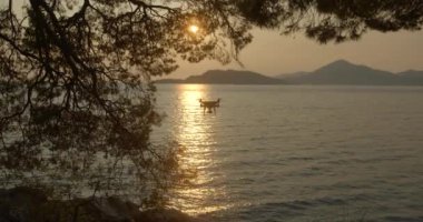 Gün batımında okyanus ve dağların üzerinde uçan helikopterdeki ağaçların arasından bak. Tekne ve katamaran suda yüzüyor. 4k.