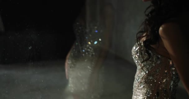 スタジオでポーズをとるモデル。暗闇の中でガラスの前にポーズをとって輝くドレスを着た美しい女性の劇的な写真 — ストック動画