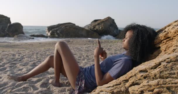 A gyönyörű afrikai lány szép mosollyal fekszik a homokon, közel a sziklához, és SMS-ezik, csevegnek és böngésznek a mobiltelefonon keresztül.. — Stock videók