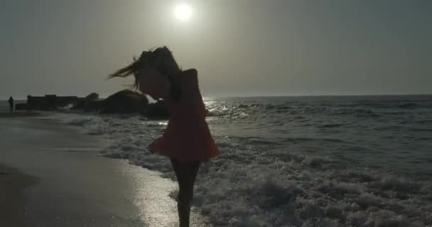 Maravillosa morena con el pelo largo en el vestido rosa es felizmente dando vueltas y ordenando el pelo mientras corre a lo largo de la costa durante la puesta del sol. — Vídeo de stock