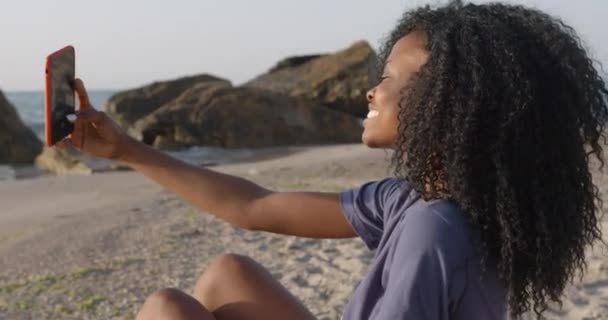 Boczny portret atrakcyjnej młodej afrykańskiej dziewczyny z ładnym uśmiechem i ciemnymi kręconymi włosami robiącej selfie przez telefon komórkowy na plaży. — Wideo stockowe