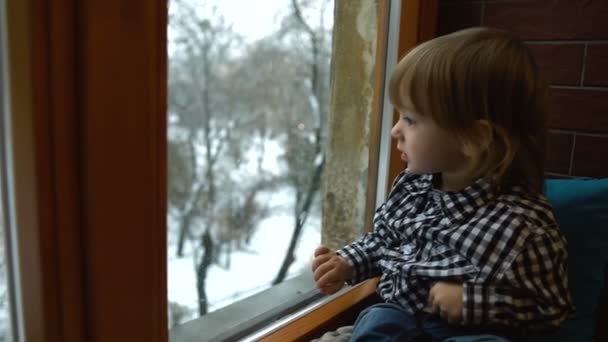 Πορτρέτο του όμορφου μικρού αγοριού με τα ξανθά μαλλιά που κάθεται στο περβάζι του παραθύρου και κοιτάζει τον χιονισμένο καιρό μέσα από το παράθυρο. — Αρχείο Βίντεο