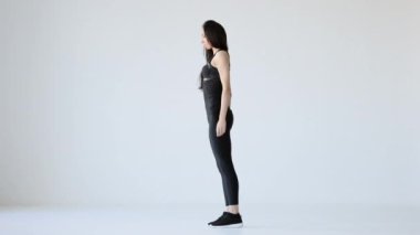 Spor giyimli sporcunun yan görüntüsü kapalı alanda geriliyor. Beyaz arka planda bir ayağının ucuna kadar eğiliyor..