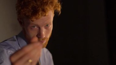 Kızıl saçlı adamın yakın plan portresi. Sakalı var. Siyah arka planda eli olan bir jest parası var..