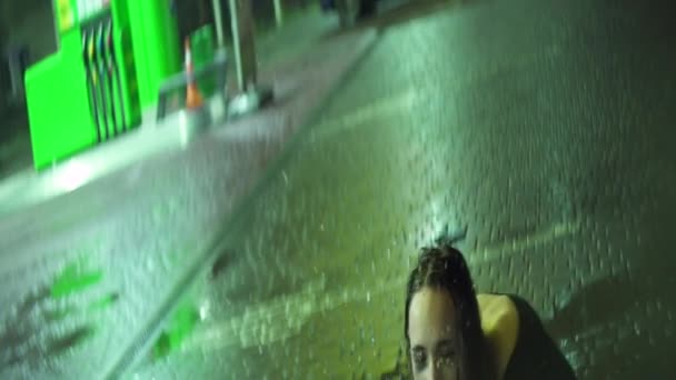 黒のドレスを着た若いブルネットの女性とスミアメイクはヒステリーにあります。彼女は夜雨の中地面に座りながら泣いている。. — ストック動画