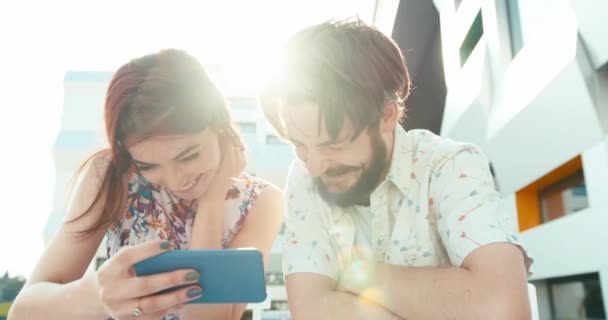 Belo casal amoroso de hipsters estão rindo enquanto assiste vídeo no telefone celular nos solários. Luz de sinalização. 4k. — Vídeo de Stock