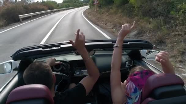 Back close-up view of the cheerful couple having fun, listening to music and waving hands while driving the cabriolet along the road in the mountains. — Stock Video