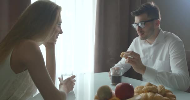 Gros plan portrait du jeune couple joyeux qui parle joyeusement pendant le petit déjeuner à la maison. Le bel homme aux lunettes boit du café avec des croissants. — Video