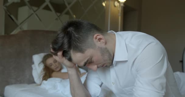 Paar mit Problemen im Bett. Der deprimierte Brunet macht sich Sorgen, während er auf dem Bett vor dem verschwommenen Hintergrund seiner wütend enttäuschten blonden Frau im Tanga sitzt, die das Zimmer verlässt.. — Stockvideo