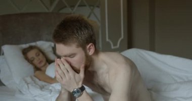Depresyondaki kızıl saçlı adam yataktaki problemler için endişeleniyor. Yatakta uzanmış bekleyen üzgün sarışın kadının bulanık arka planı..