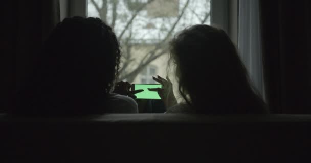 Ξανθιά και μελαχρινή κοπέλα φίλοι μιλούν και βλέποντας κάτι στο κινητό τηλέφωνο με πράσινη οθόνη, ενώ κάθεται στον καναπέ. Πίσω όψη. Τοποθεσία κατοικίας. Χωρίς πρόσωπο. — Αρχείο Βίντεο