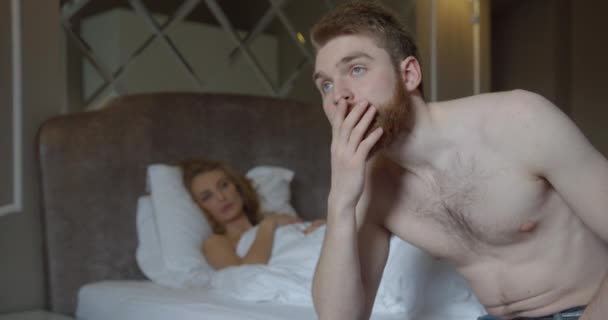 Közelkép a depressziós vörös hajú férfiról, aki az ágyon ül a szőke felesége homályos hátterében, aki rajta fekszik.. — Stock videók