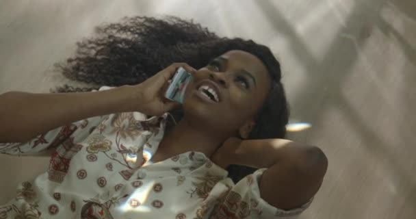 Charmante brune africaine heureuse avec maquillage naturel rit et parle via le téléphone portable tout en étant couchée sur le sol. Portrait supérieur. — Video