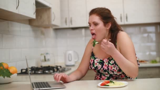 食事中の太りすぎの女性は、ラップトップを介してネットをチャットし、サーフィンしながら笑っていると台所でキュウリ、トマト、パセリとマッシュポテトを食べる. — ストック動画