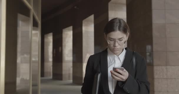 Succesvolle zakenvrouw in bril en modern pak is chatten en browsen op de mobiele telefoon tijdens het lopen naar de camera. 4k. — Stockvideo
