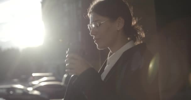 Retrato lateral de la joven empresaria de éxito en traje y anteojos disfrutando del café paseando bajo los rayos del sol. Imágenes de 4k. Luces de destello. — Vídeo de stock