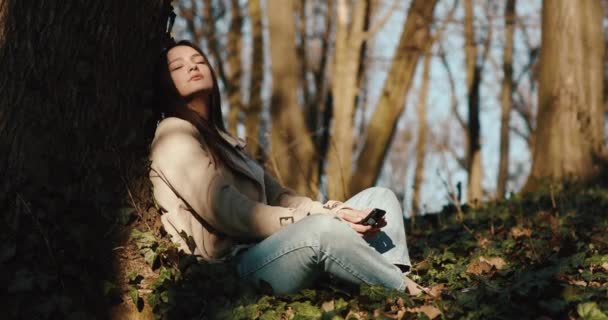 秋の波。女性は公園の芝生の上に座っている朝の太陽の光を楽しんで、彼女の腕の中でスマートフォンを持っています — ストック動画