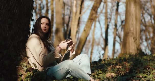 Sonbahar titreşimleri. Parktaki çimlerde oturan ve kollarında akıllı bir telefonla sabah güneşinin tadını çıkaran bir kadın. — Stok video