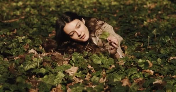 Mulher pensativa e sonhadora encontra-se nas folhas caídas no parque e olha para longe — Vídeo de Stock